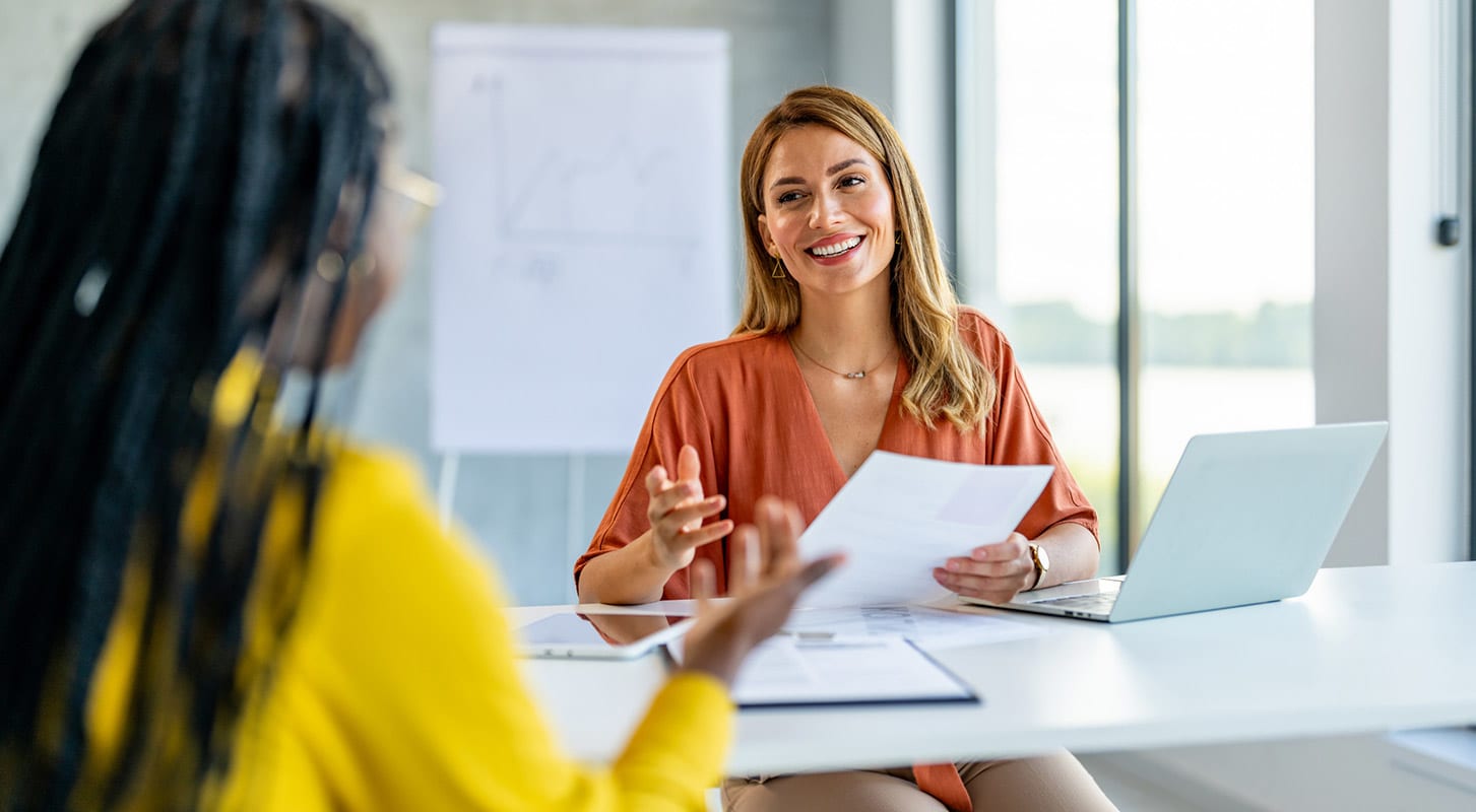 Two women are seated at an office table engaged in a conversation.